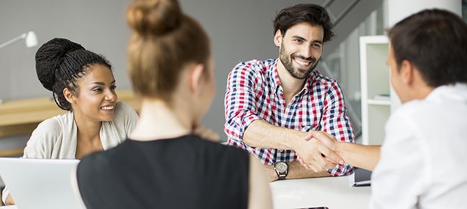 a group of people in a meeting look over at two men shaking hands.
