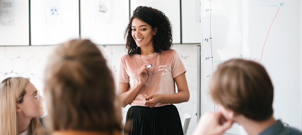 A young woman stands in front of a whiteboard during a meeting.